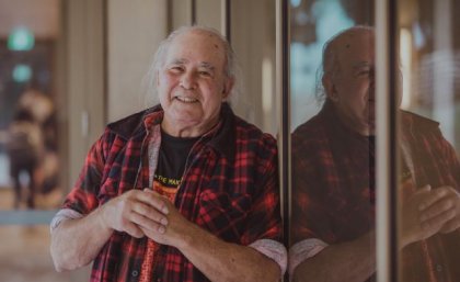 a man in a check shirt leans against a window in which he is reflected